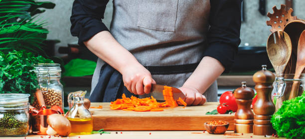 istock Chef food blogger cuts carrots into strips. Cozy kitchen with wooden table, kitchenware, vegetables, herbs and ingredients for cooking. Healthy vegan food, culinary, recipes, food blogging 2205621179