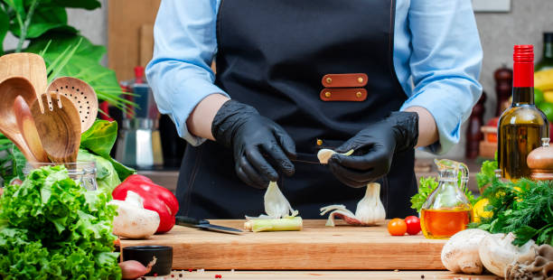 istock Chef in gloves peels young garlic. Cozy kitchen with wooden table, kitchenware, vegetables, herbs and ingredients for cooking. Healthy vegan eating, culinary, recipes, food blogging 2205621189
