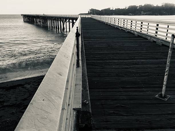 A black-and-white photo of the pier at San Simeon, California, extending over the ocean with a distant shoreline.