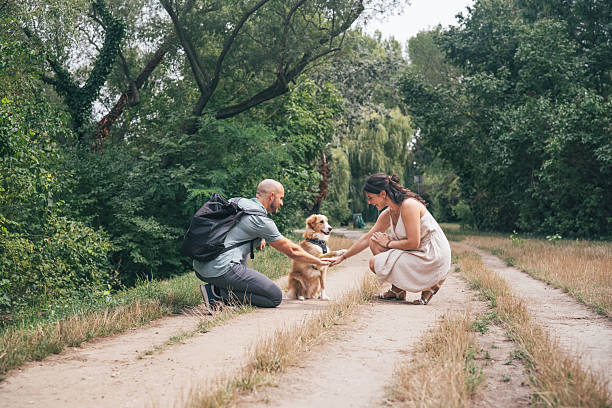 A cheerful couple interacts with their adorable dog on a scenic dirt path surrounded by lush greenery. The woman and man kneel down, joyfully holding the dog's paws, sharing a warm and loving connection. The peaceful natural setting enhances the charm of this heartfelt moment