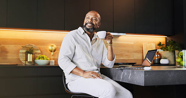 Thinking, home and black man in kitchen, coffee and smile with laptop, ideas and problem solving. African person, apartment and entrepreneur with herbal tea, morning and daydreaming with solution