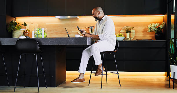 istock Remote work, phone and black man in kitchen with laptop for research, online networking and website. Professional, home and person with smartphone for finance consulting, insurance and planning 2205649041