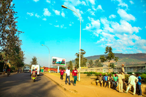 A vibrant street scene in Kigali, Rwanda, featuring pedestrians, motorcyclists, and a group of people gathered near the roadside. The image captures the daily rhythm of urban life against the backdrop of Kigali’s hilly landscape and bright blue sky. This editorial image showcases the city’s infrastructure, social interactions, and transport culture.