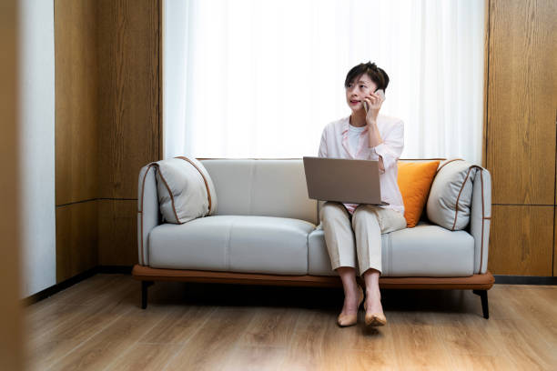 istock Woman sitting on sofa using laptop and phone 2205655422