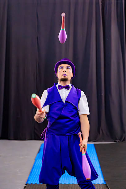 Juggler wearing a purple costume and hat skillfully juggles colorful clubs on a stage with a black backdrop