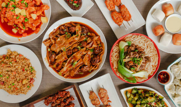 A close-up captures a variety of vibrant Chinese dishes laid out on a wooden dining table, including spicy diced chicken and saucy eggplant. Hands holding chopsticks are prominent, reaching for different dishes, conveying a communal and authentic Chinese dining experience.