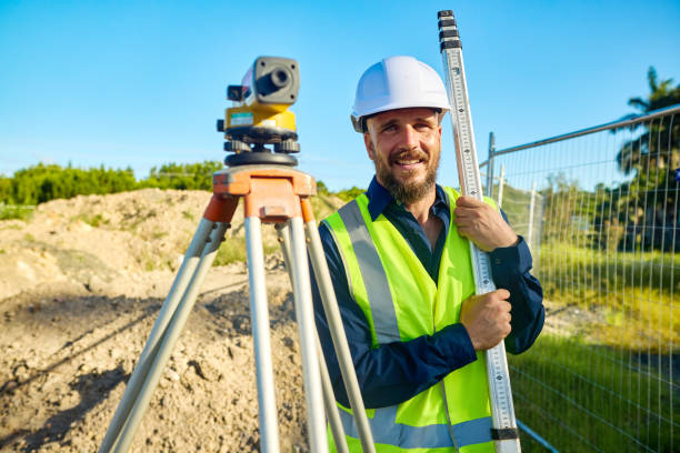 Surveyor smiling and holding a staff next to a theodolite, working on a construction site