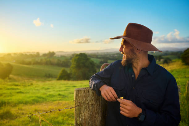 Australian farmer smiles while leaning on a fence post, enjoying the sunset over his green, rolling hills