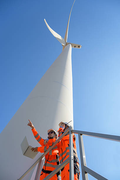 istock Two engineers in safety uniform inspecting wind turbine. Male worker holding laptop while female communicates via radio walkie talkie while standing on platform 2205666796