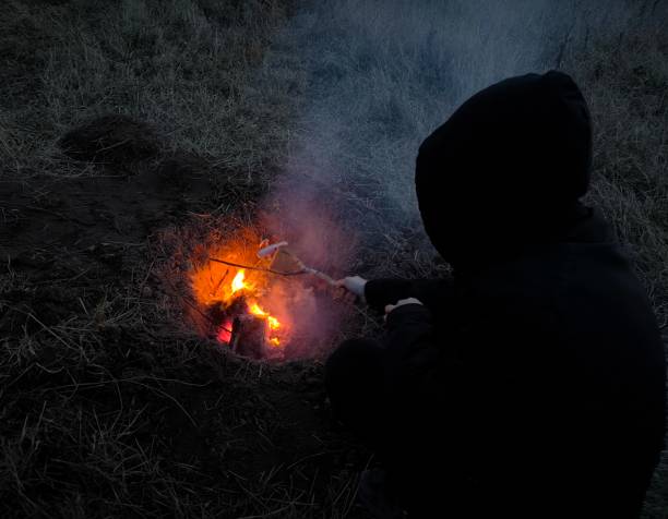 istock Back view, guy is sitting alone by a campfire on a dark night. Concept of camping. 2205683549
