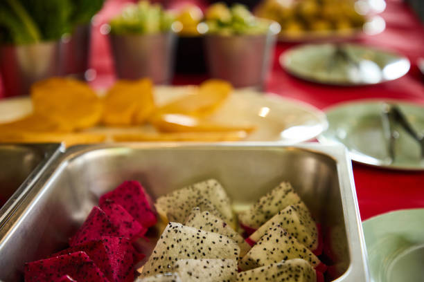 Slices of vibrant dragon fruit placed in a serving tray, surrounded by a blurred background of various dishes, conveying a fresh and colorful buffet or catering presentation.