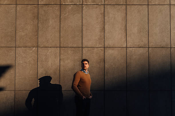 Young creative entrepreneur standing with hands in pockets in front of a modern building facade, projecting a long shadow