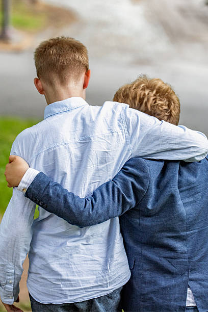Two boys, seen from behind, walking side by side on a grassy hill, holding each other in a supportive embrace