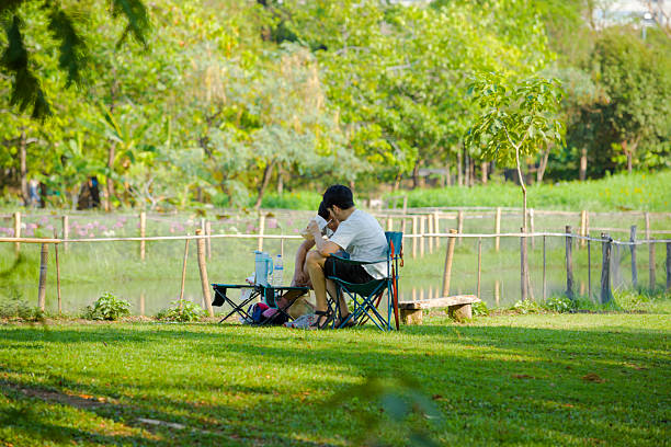 Two young thau adults are having a picnic in Wachirabenchathat Park (Rot Fai Park) in Bangkok Chatuchak. They are wearing casual clothes and are sitting on foldable chairs/seats. In front of them is a foldable small table. One person has eywear and pink socks. Man in foreground is wearing sandals. In background is one of the lakes in the park.