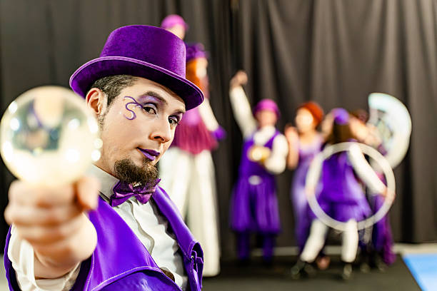 Magician with purple costume and make-up holding a crystal ball, performing a magic trick with circus performers in the background