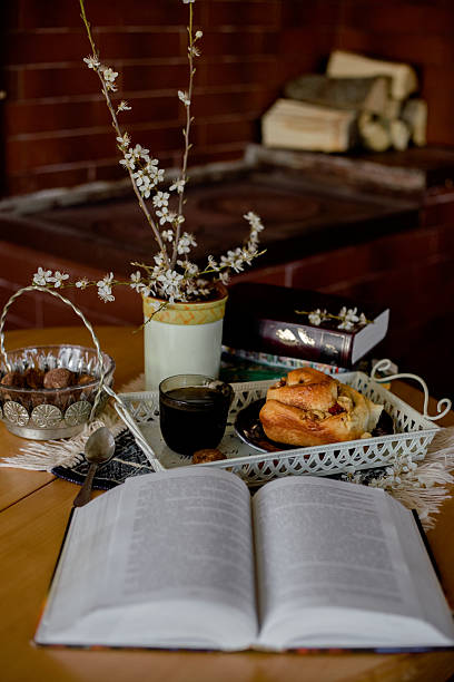 istock Cozy reading nook: open book, coffee cup and fresh pastries on a wooden table by a brick stove 2205700655