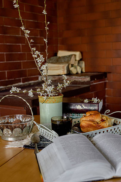 istock Cozy reading nook: open book, coffee cup and fresh pastries on a wooden table by a brick stove 2205700657