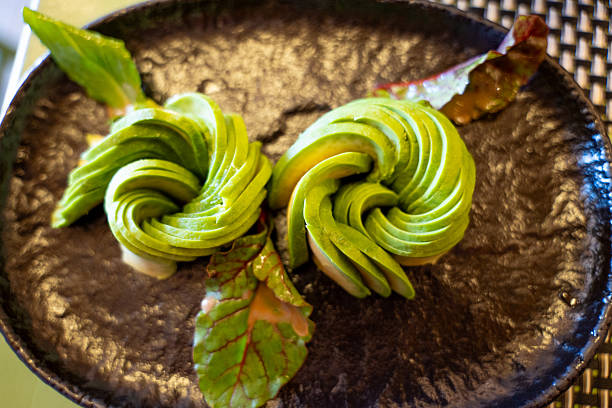 Intricately carved avocado slices arranged as rose flowers on a dark ceramic plate. Fresh and artistic food presentation. Ideal for health-food and culinary art concepts.