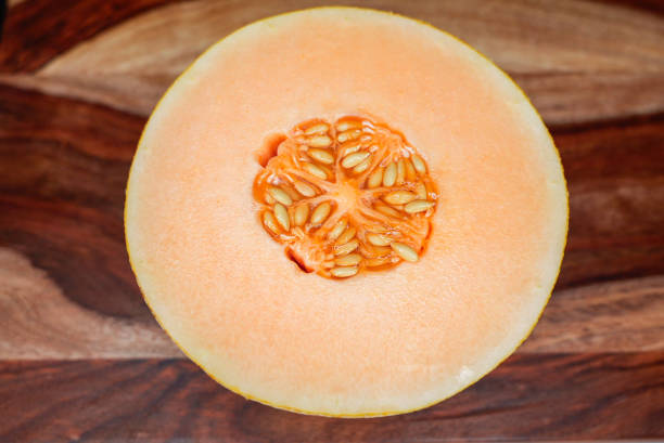 A ripe cantaloupe melon, one half cut open revealing its juicy orange flesh and seeds, placed on a natural wooden surface, showcasing its vibrant texture and freshness.