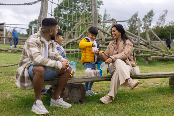 Wide view of a family including two young brothers, a mother and a father at an outdoor adventure playground at a Zoo in Northumberland, North East England. They are sitting on a wooden bench, taking a break from playing to enjoy a picnic.Videos similar to this scenario are available.