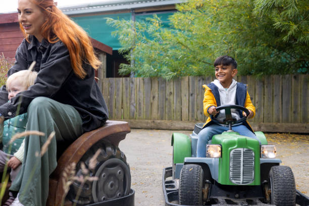 Wide view of a young girl and mother playing and driving on a coin-operated mini tractor go-kart at an outdoor adventure playground at a Zoo in Northumberland, North East England. A young boy is riding behind them.\n\nVideos similar to this scenario are available.