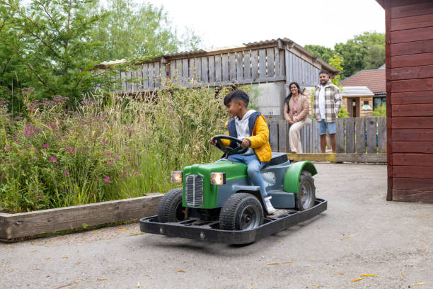 Wide view of a young boy playing and driving on a coin-operated mini tractor go-kart at an outdoor adventure playground at a Zoo in Northumberland, North East England. His mother and father are watching him from the sideline.