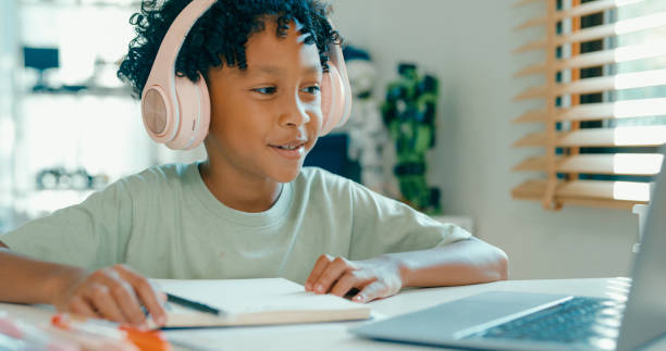 istock Young African-American boy wearing headphones, is deeply focused while participating in online learning session from home. Education courses online. 2205736403