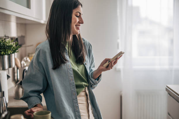 istock Smiling woman using smartphone and drinking coffee in kitchen 2205746330
