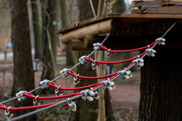 A sturdy rope bridge made with red ropes and wooden supports connects a treehouse to the ground. Surrounding trees provide a natural backdrop, creating an adventurous atmosphere.