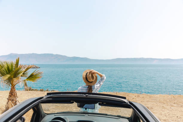 A woman standing in a convertible car enjoys the view of the ocean on a sunny summer day, exemplifying relaxation, travel, and the spirit of adventure.