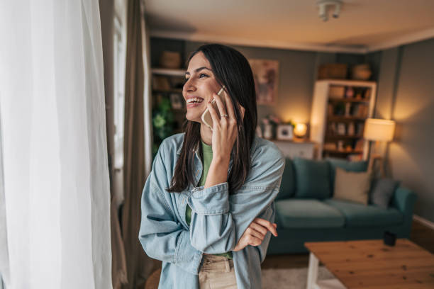 istock Young woman talking on mobile phone and looking through window at home 2205765901