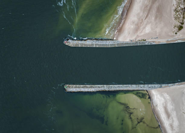 An aerial view shows a river mouth where dark water flows between two stone jetties into the sea, with sandy beaches on either side.