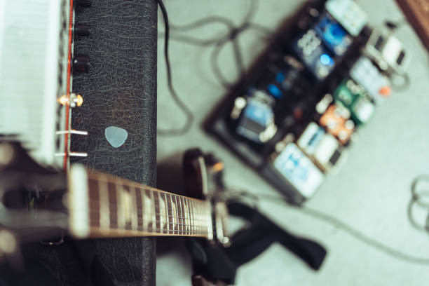 Electric guitar resting on an amplifier, with a guitar pick placed on top. A pedalboard with various effect pedals is visible in the background.