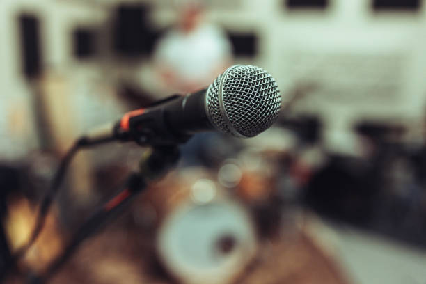 Close-up of a microphone in a rehearsal studio, with a blurred drum set in the background. The image captures the essence of live music and performance.