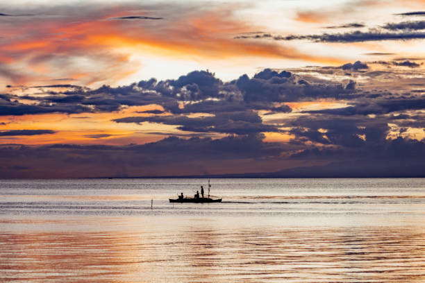 A serene view of fishermen in silhouette navigating a boat across calm waters during a vivid sunset in Siquijor, Philippines, with vibrant skies enhancing the captivating moment.