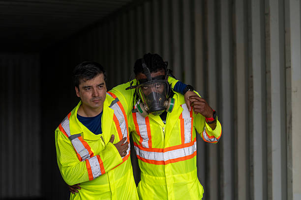 Two industrial workers in high-visibility suits assist each other after a hazardous incident near a shipping container. Emphasizing occupational safety, teamwork, emergency response, and risk management.