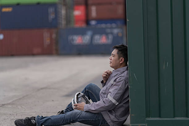 An exhausted shipping yard worker takes a break, leaning against a cargo container while holding a safety helmet. The image captures the challenges of working in global freight and logistics.