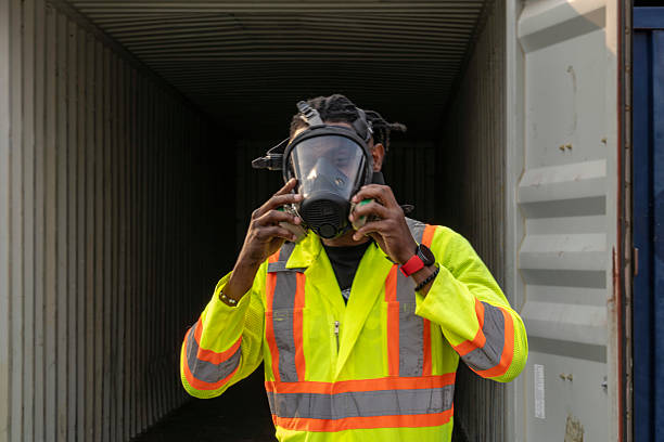 A worker in high visibility safety gear puts on a protective gas mask while standing at the entrance of a shipping container. Emphasizing industrial safety, hazardous environments, and workplace protection.