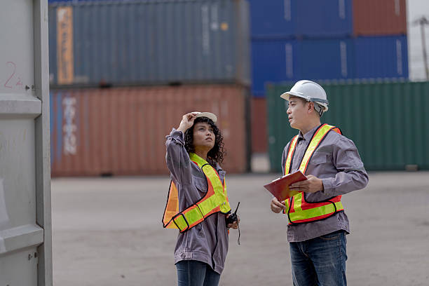 istock Two logistics workers asian man and woman discuss operations at a busy shipping container yard. One is holding a tablet while pointing towards the cargo stacks. 2205779497