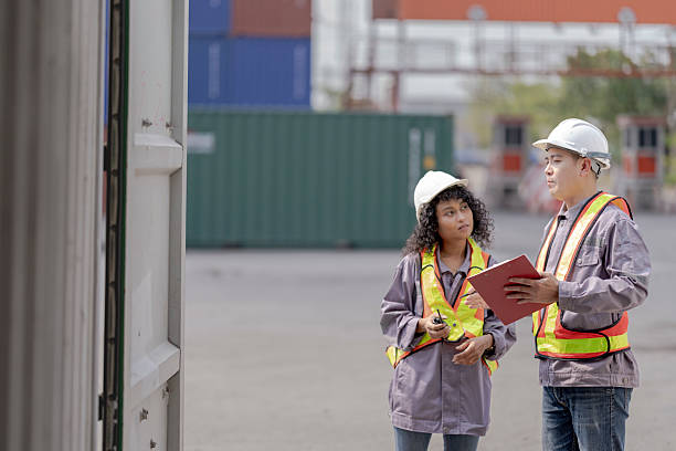 istock Two logistics workers asian man and woman discuss operations at a busy shipping container yard. One is holding a tablet while pointing towards the cargo stacks. 2205779631