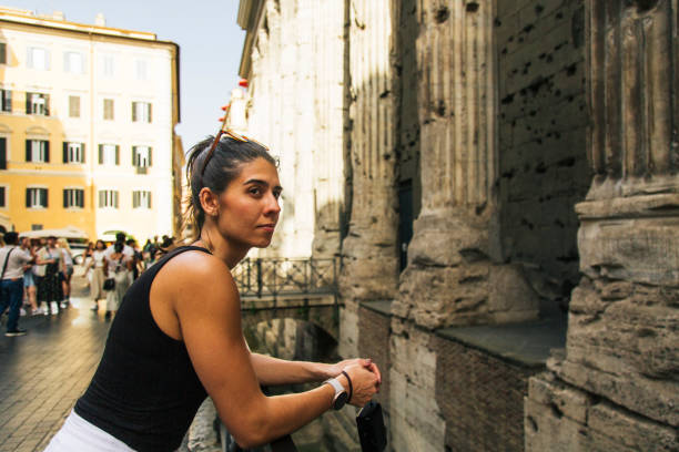 A woman stands thoughtfully in front of ancient Roman columns, appreciating their historic architecture and intricate details. The image captures a serene moment of cultural exploration and historical admiration. Temple of Hadrian, Rome.