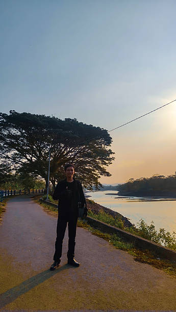 a man dressed in all black walks along a riverside road at sunset, giving a thumbs-up gesture. the golden hour light creates a peaceful and serene atmosphere, highlighting his silhouette against the glowing sky. a large, lush tree on the roadside adds depth to the composition, while the calm river reflects the warm hues of the evening. this scene captures a moment of relaxation, positivity, and appreciation for nature in a tranquil countryside setting.