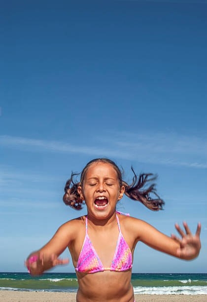 Young girl with pigtails wearing a pink bikini is shouting with excitement while playing on a sandy beach, the clear blue sky providing a vibrant backdrop to her joyful expression