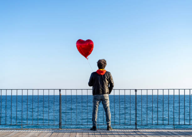 istock Lonely man holding a red heart shape balloon on the beach 2205804274