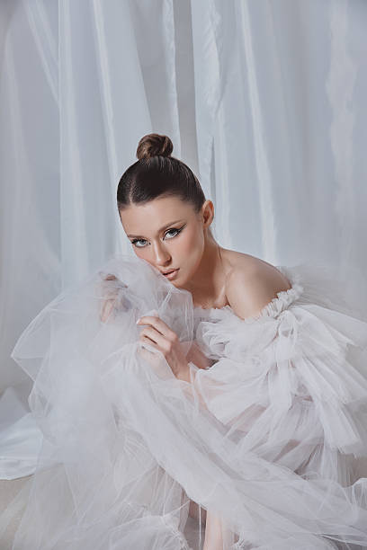 istock model showcases a delicate white tulle outfit, striking a thoughtful pose with a soft expression. serene indoor background enhances the ethereal atmosphere created by the fabric 2205820318