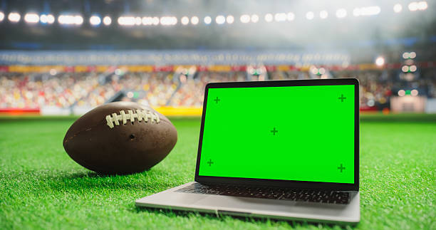 istock Laptop Computer with a Green Screen Mock-Up Display Positioned Next to American Football on a Stadium Pitch at Night. Template for Software, Streaming, Sports Betting, Multimedia Content 2205822904