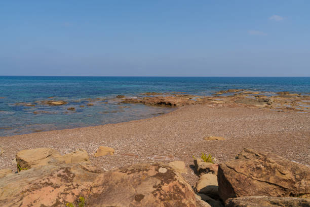 A serene view of a Mediterranean beach with a rocky shoreline and crystal-clear blue waters, under a bright blue sky, offering a tranquil coastal escape