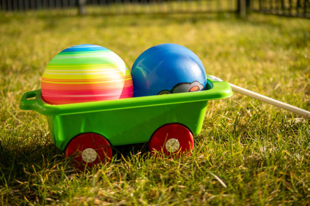 A green plastic toy wagon with two colorful balls inside, resting on sunlit grass in an outdoor setting. A bright and cheerful representation of childhood fun, play, and outdoor recreation.