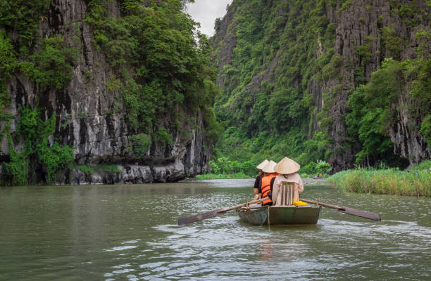 People on a boat floating down a river surrounded by tropical lush forests and mountains. Located in Tam Coc Ninh Binh Vietnam