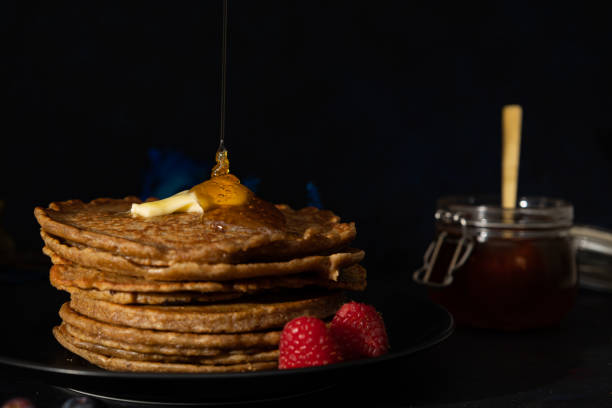 istock Front view of honey falling on stacked pancakes made with whole wheat flour with melted butter on top and raspberries on the side on a black plate on a dark background 2205828995